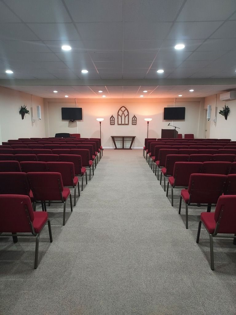 Small chapel or auditorium with rows of red chairs facing a front stage and altar-like table
