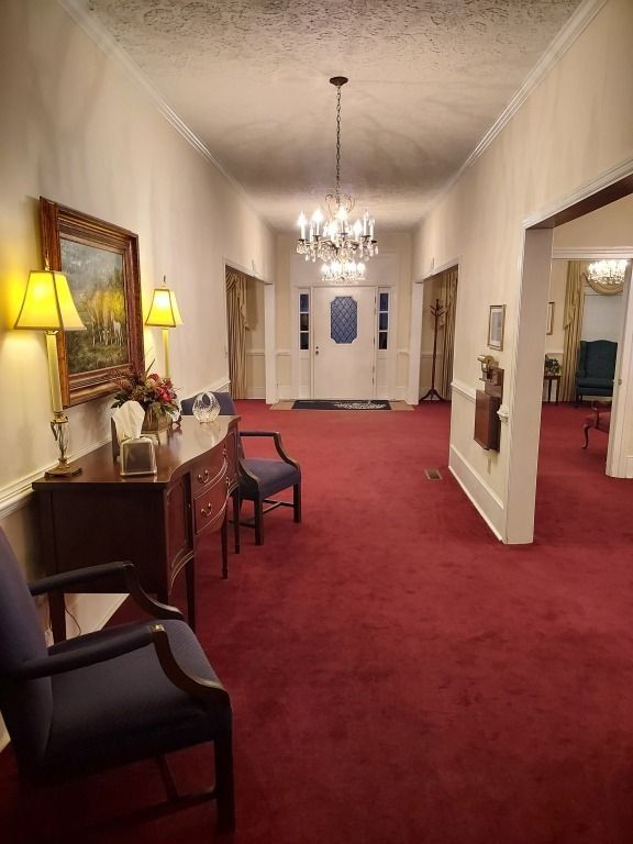 Red-carpeted hotel hallway with lamps, console table, chairs, and a chandelier at the far end