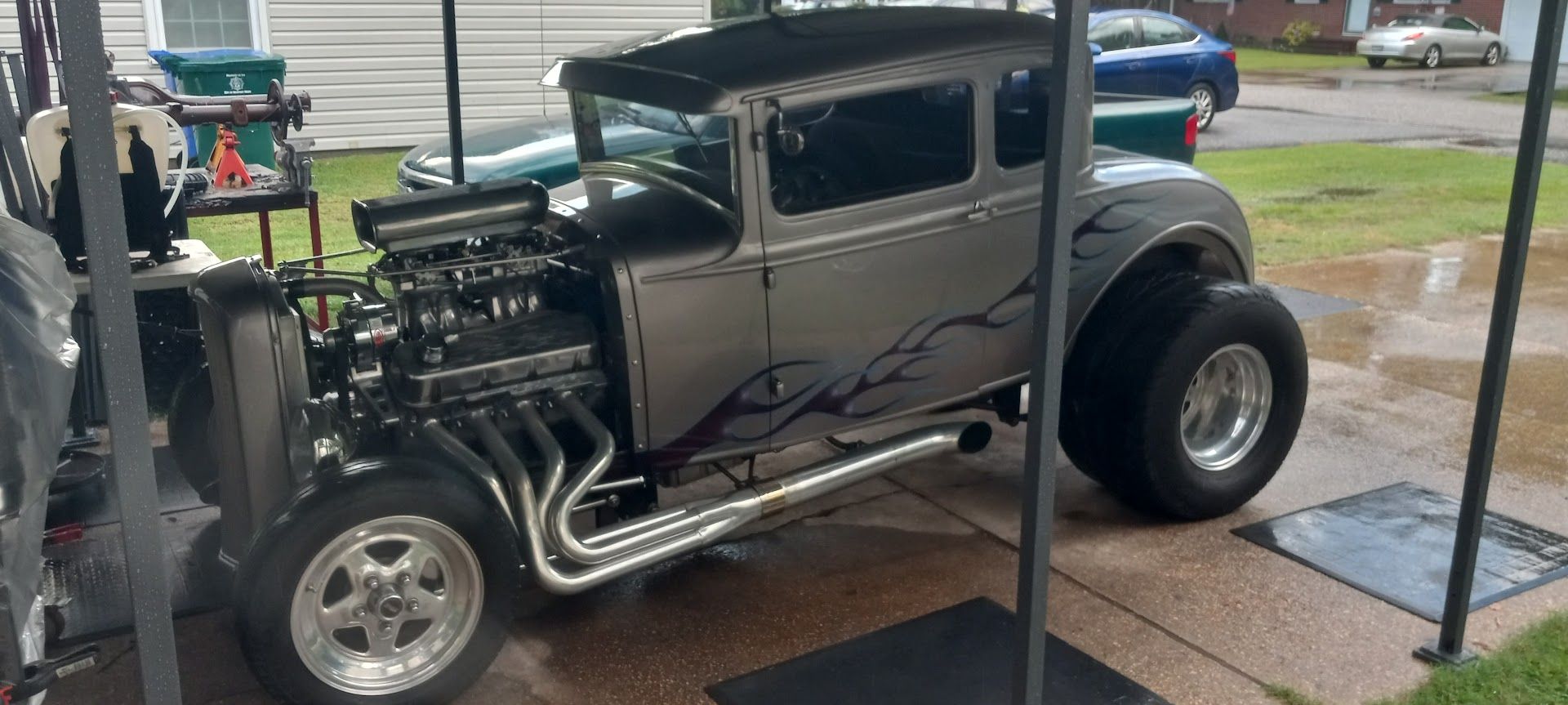 A custom silver hot rod with large rear tires, an exposed engine, and black flame decals parked outdoors. | Sprinkle and Williams II, Inc.