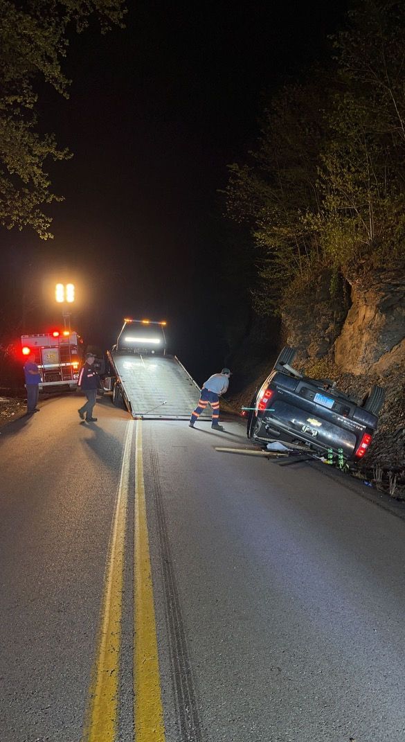 Nighttime crash scene: car off-road in ditch beside tree, with emergency vehicles and headlights on a rural road.