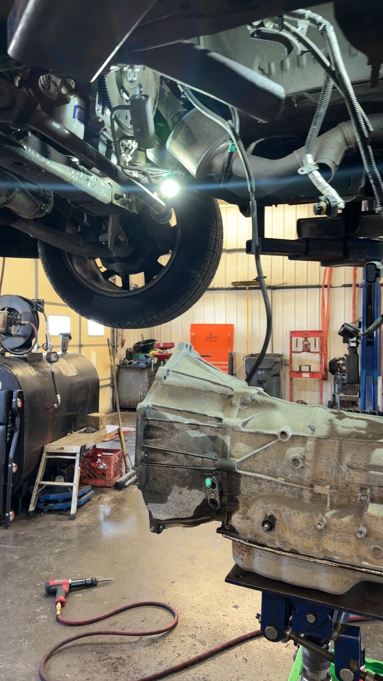 Car lifted in an auto shop, with a transmission on a stand and tools on the floor.