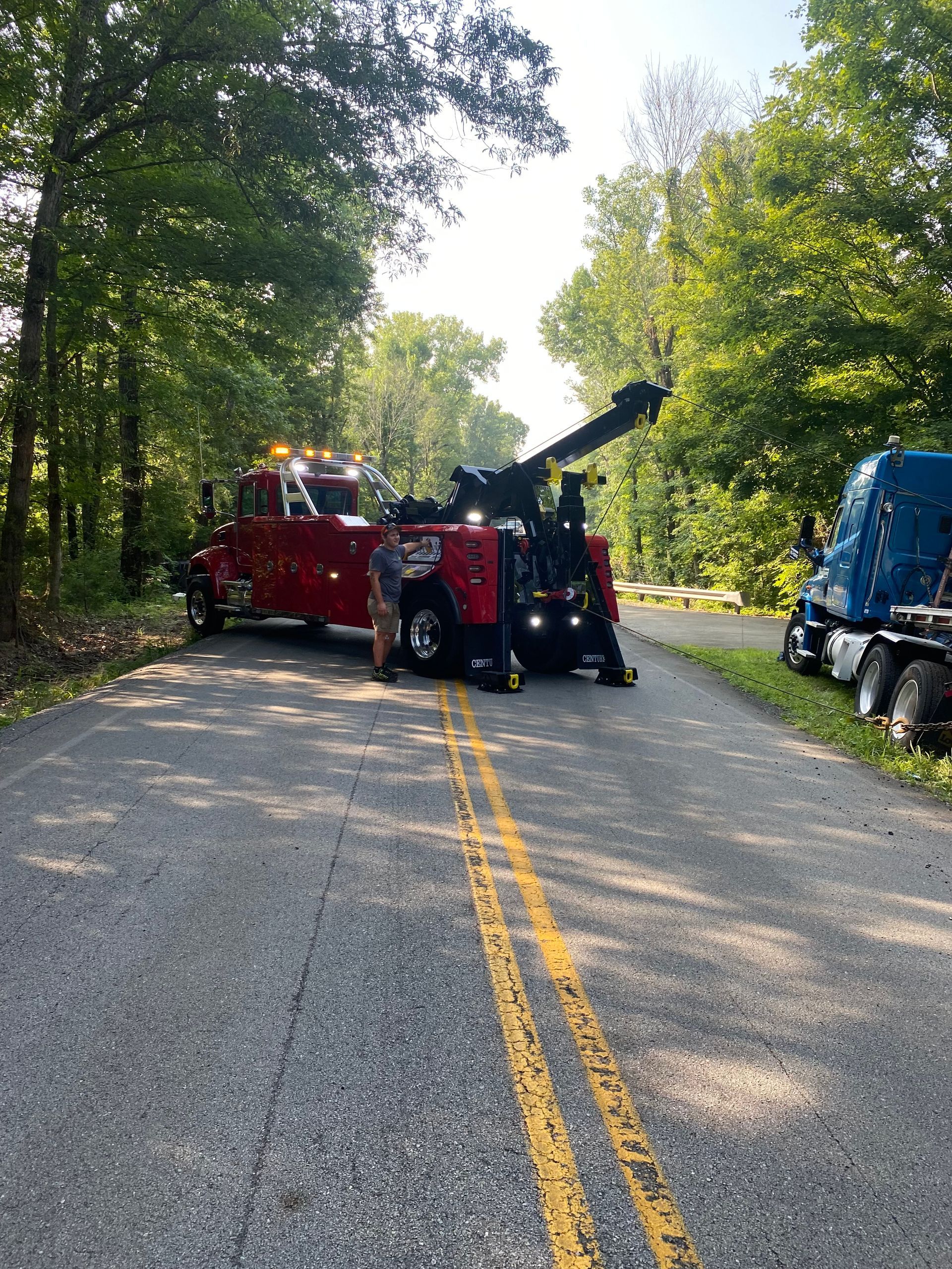 Red tow truck blocking a tree-lined road with a blue truck parked at the right shoulder