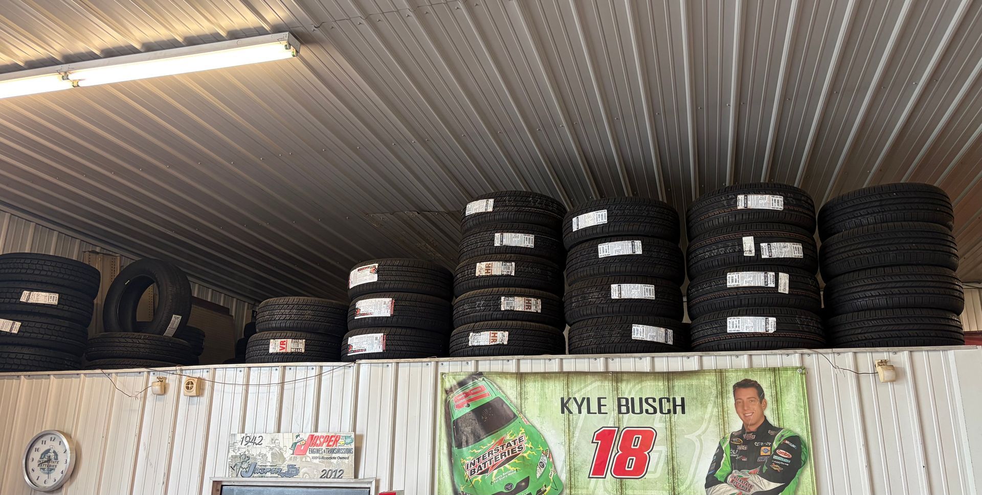Stacks of black tires stored on shelves in a garage, with racing posters on the wall below.