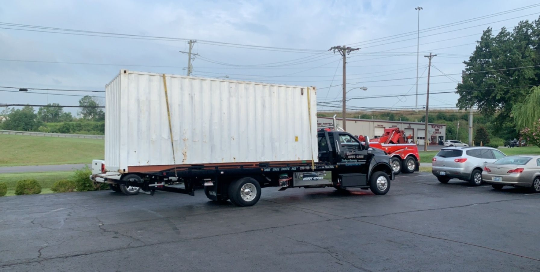 Tow truck hauling a white box truck on a roadside near parked cars and power lines
