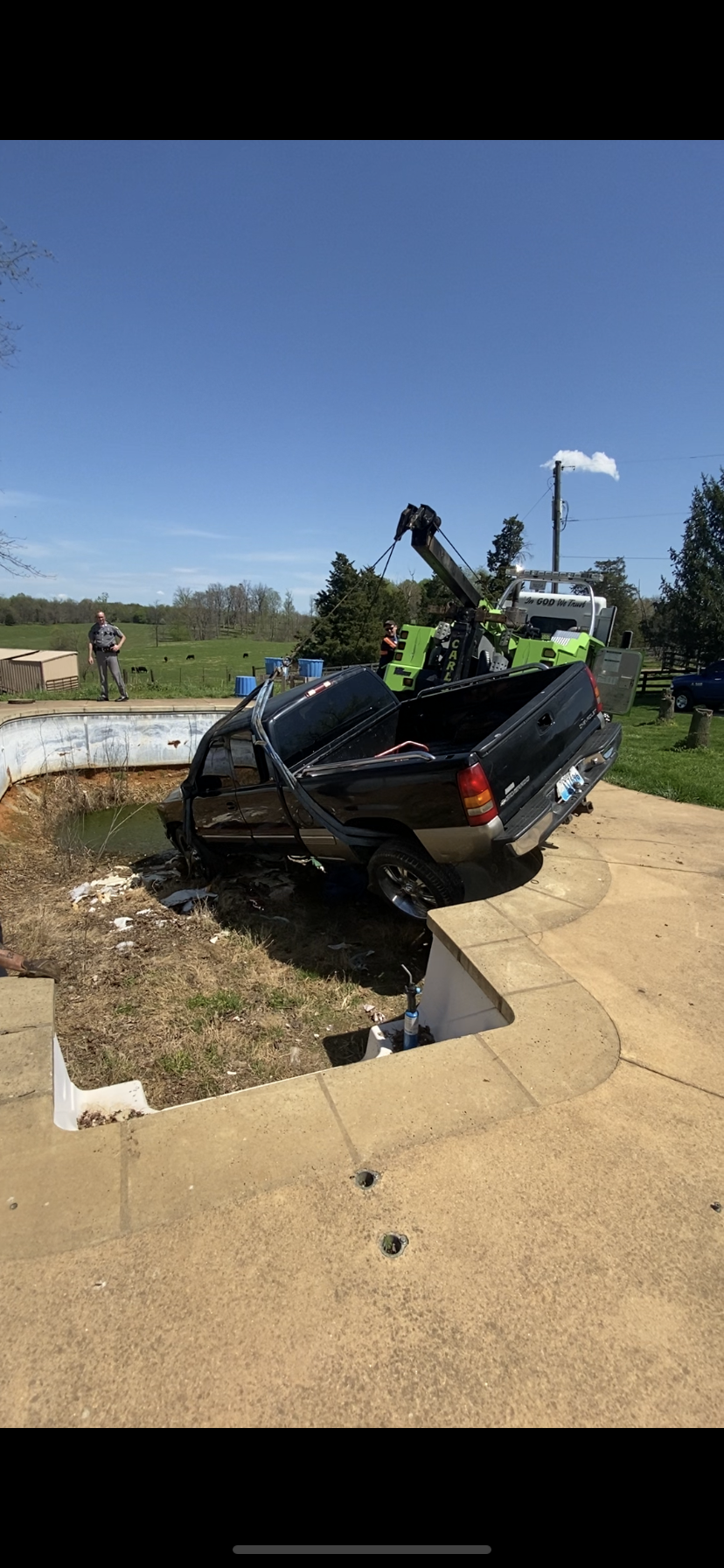 Black car crashed into a ditch beside a sidewalk, with an open hood and tow truck equipment nearby