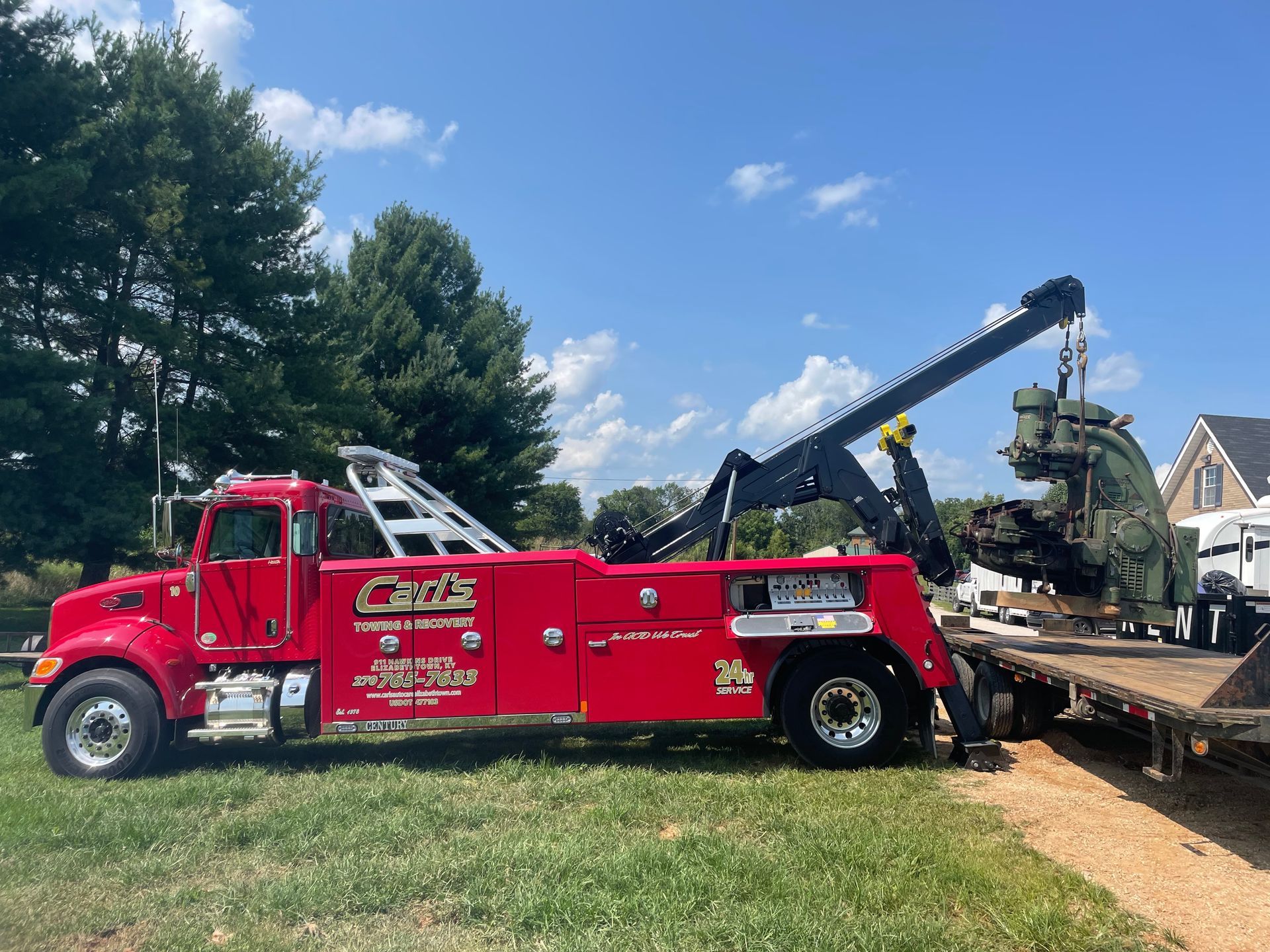 Red tow truck loading a damaged vehicle on a flatbed beside trees under a blue sky