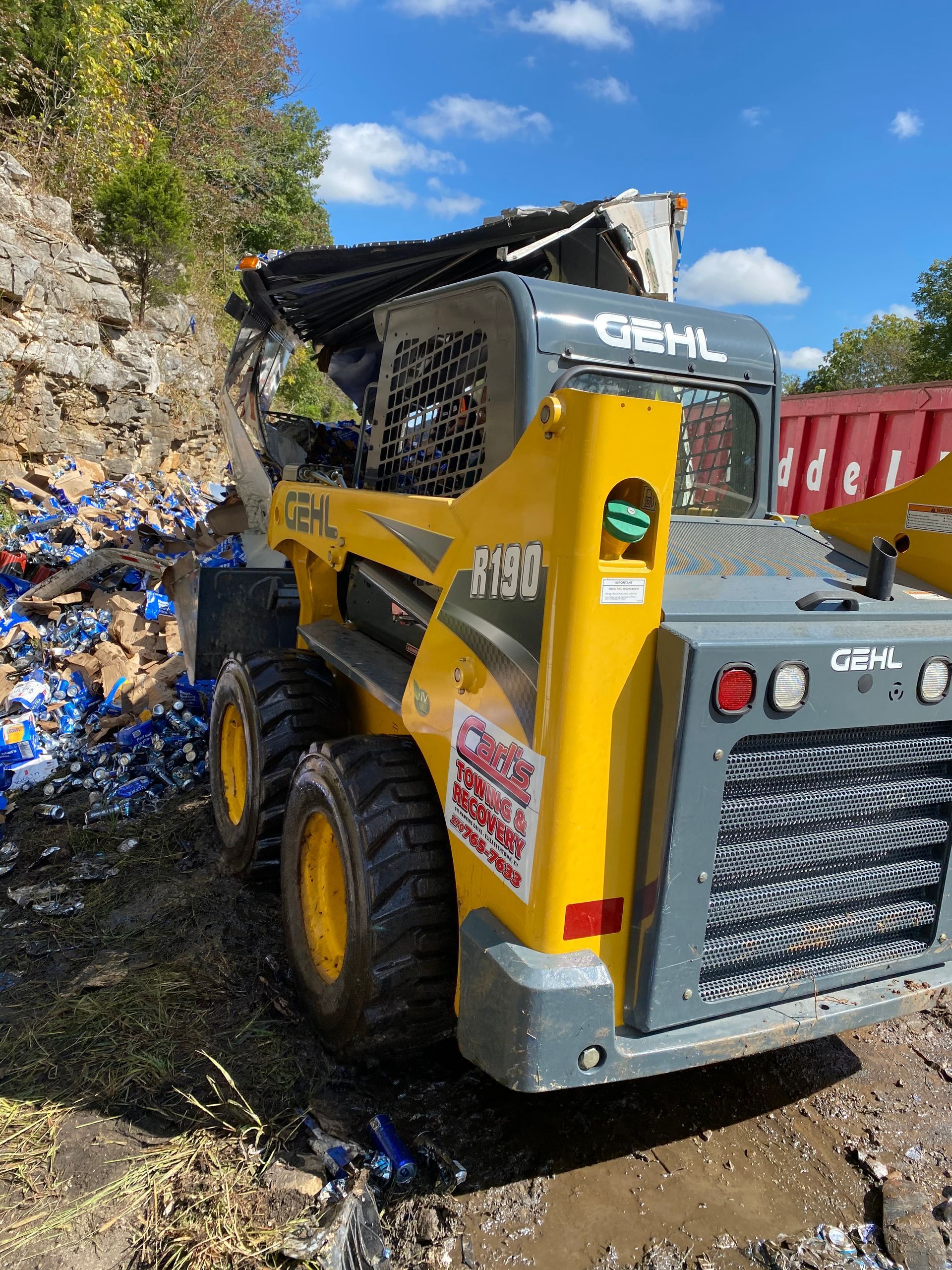 Yellow Gehl skid-steer loader on a muddy path beside a rock wall and red container on a sunny day
