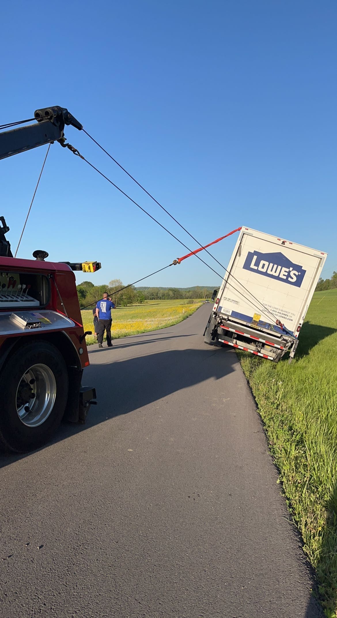 Tow truck pulling a tipped white box truck off a rural road under a clear blue sky