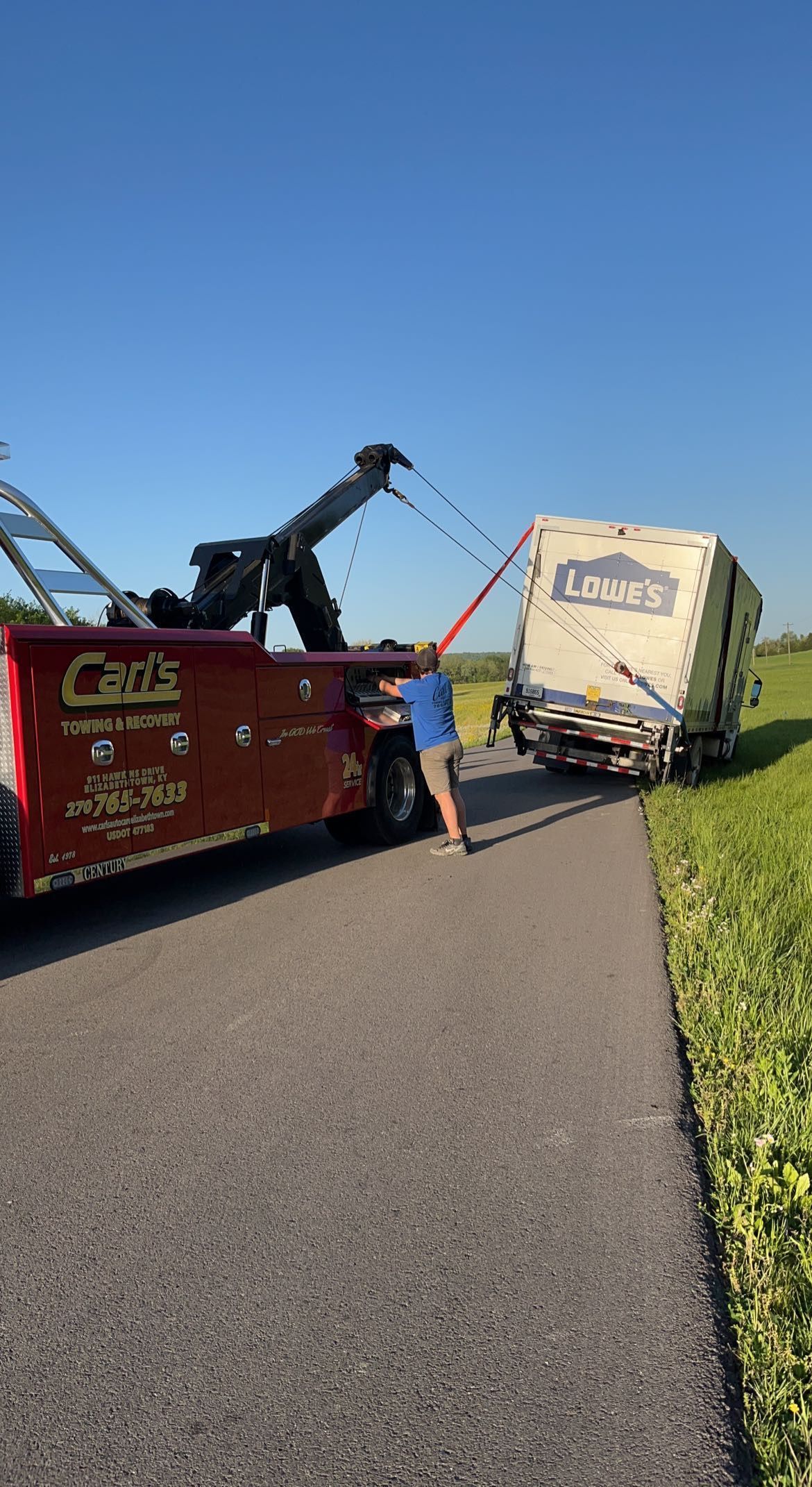 Tow truck lifting a tipped white trailer on a rural road beside a grassy field under a clear blue sky