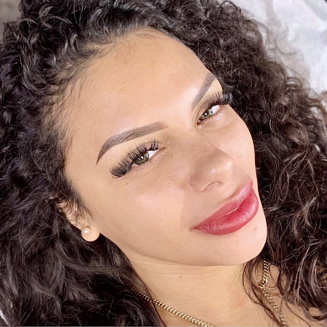 Woman with curly hair, makeup: dark eyebrows, long lashes, red lips. Smiling, lying down.