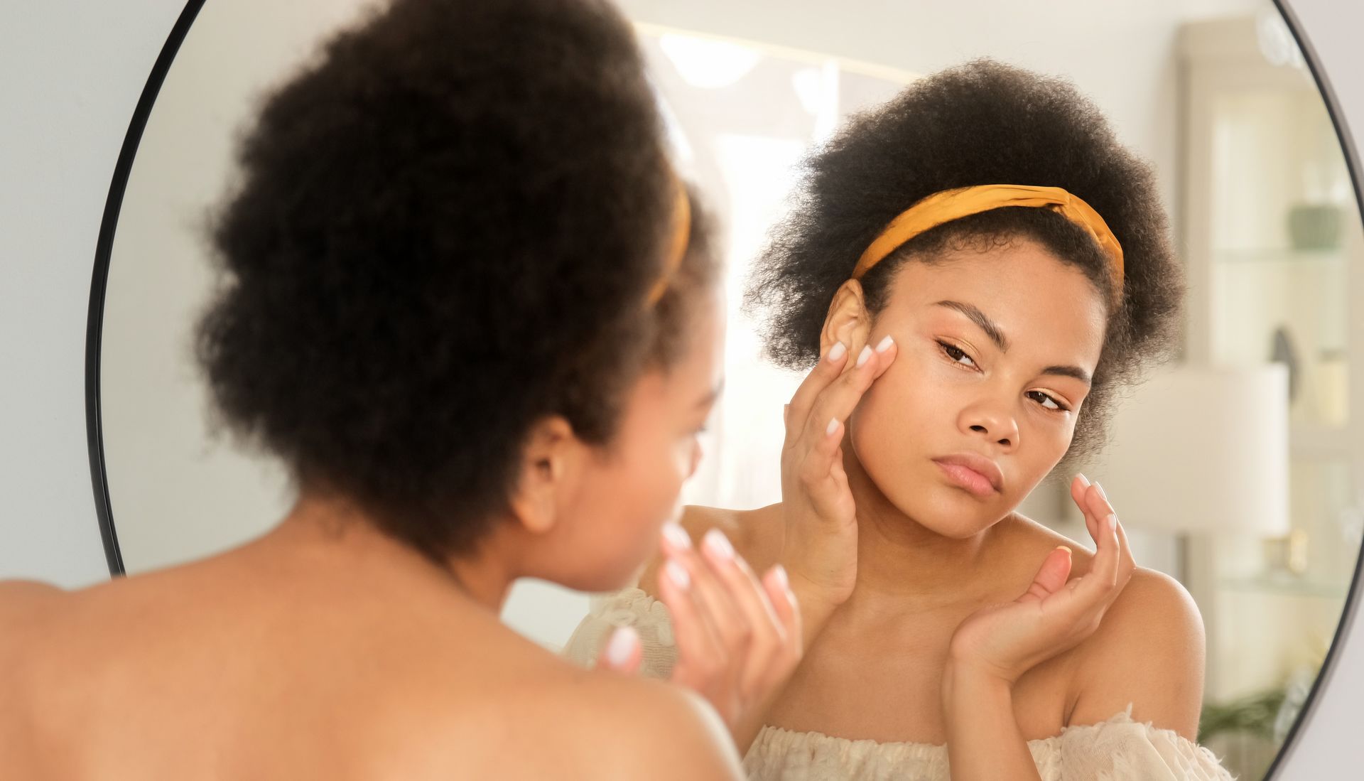 Woman looking in a mirror, touching her face. She wears a yellow headband; the mirror is round.
