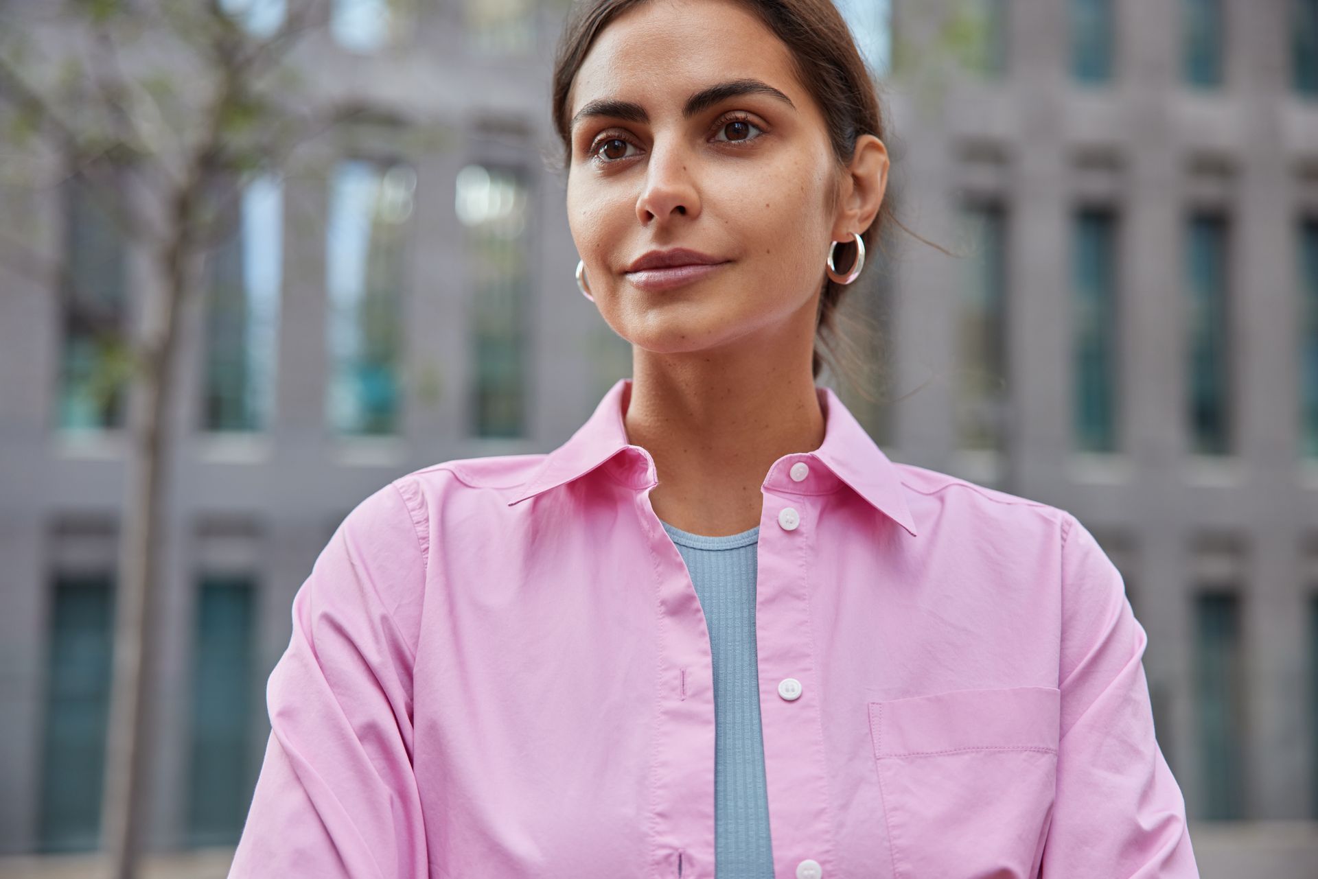 Woman in pink shirt outdoors, looking toward the side with neutral expression, silver hoop earrings, building in background.