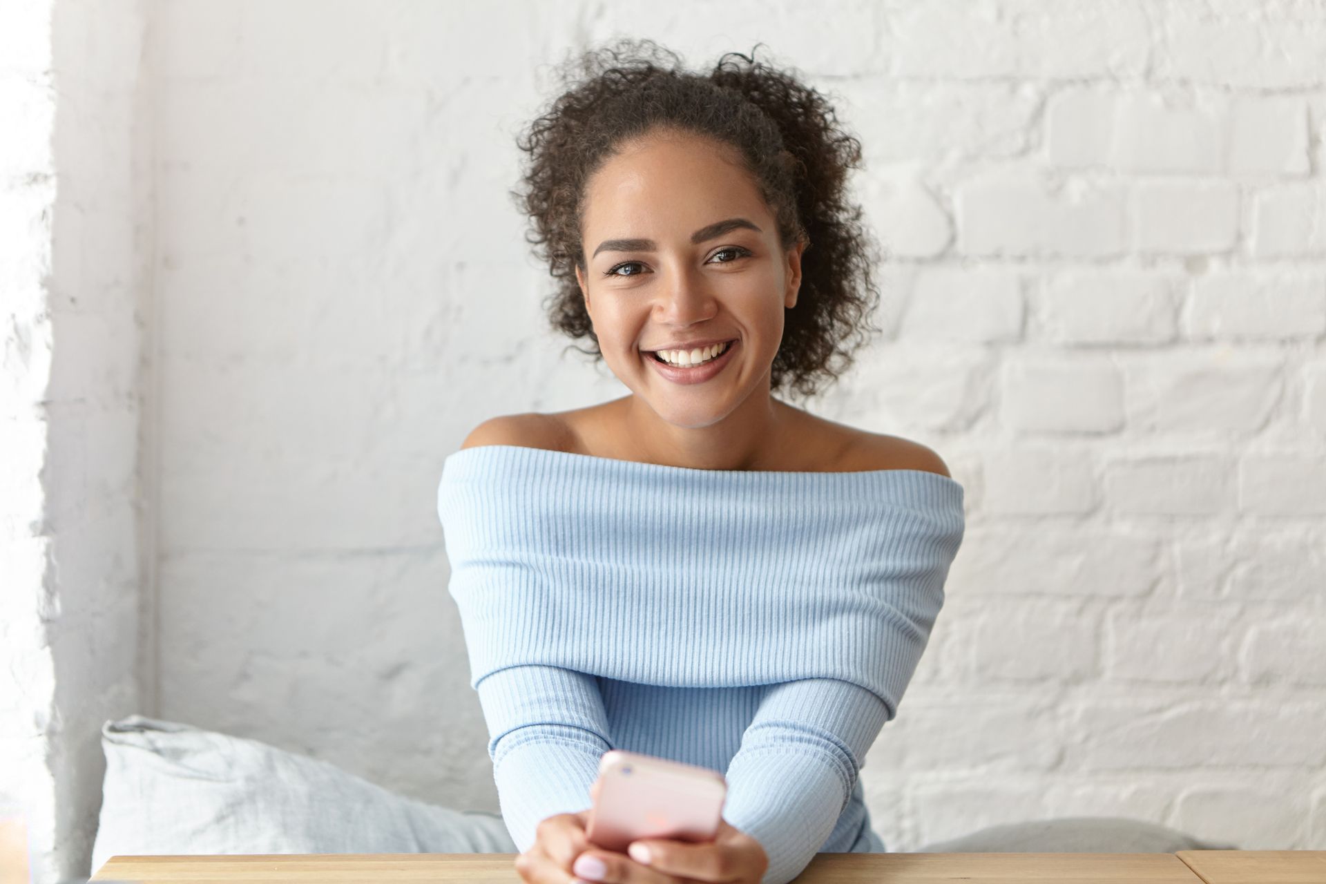Woman smiling, wearing a light blue off-the-shoulder sweater, holding a phone, sitting in front of a white brick wall.