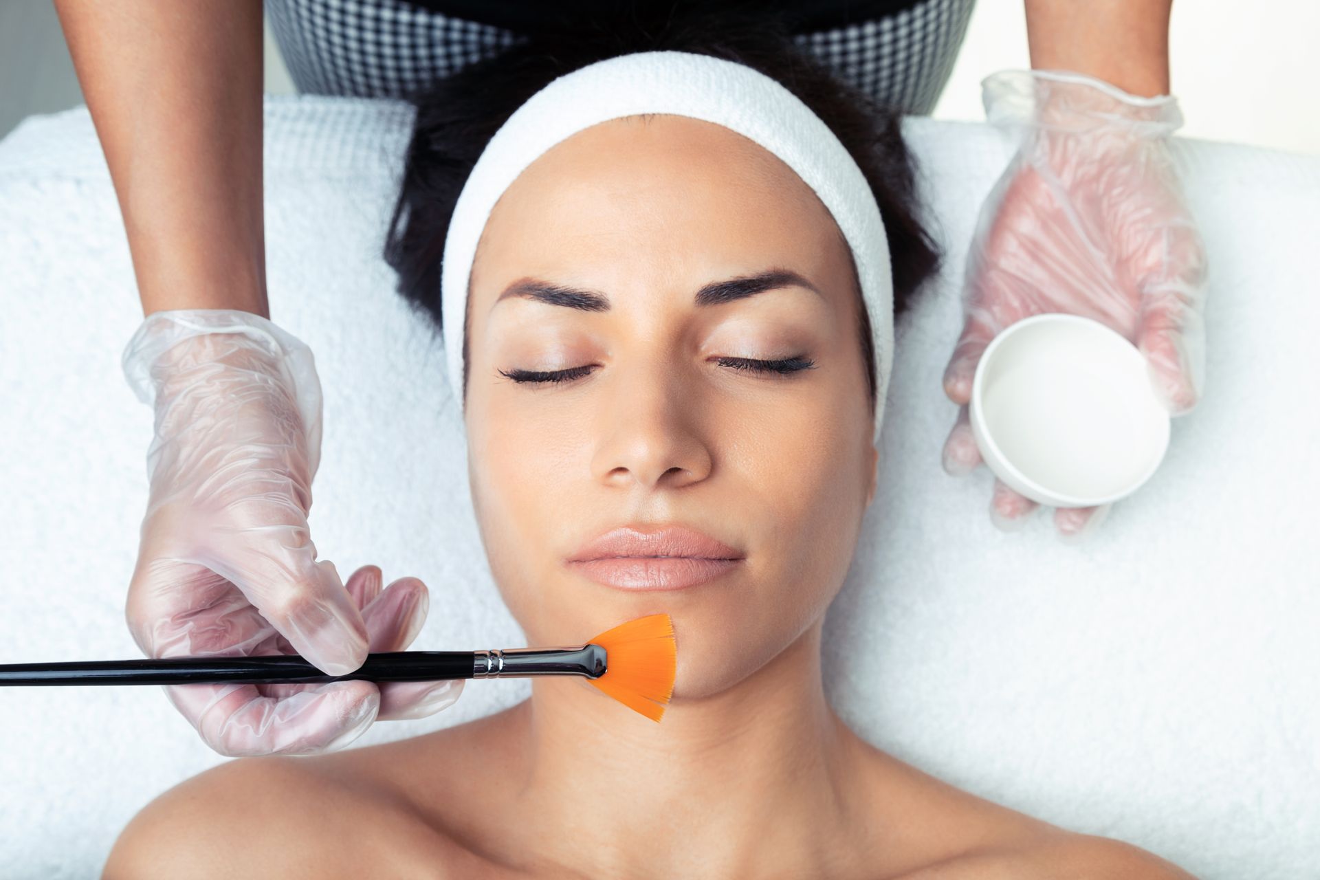 Woman receiving facial treatment, hands in gloves holding brush and bowl.