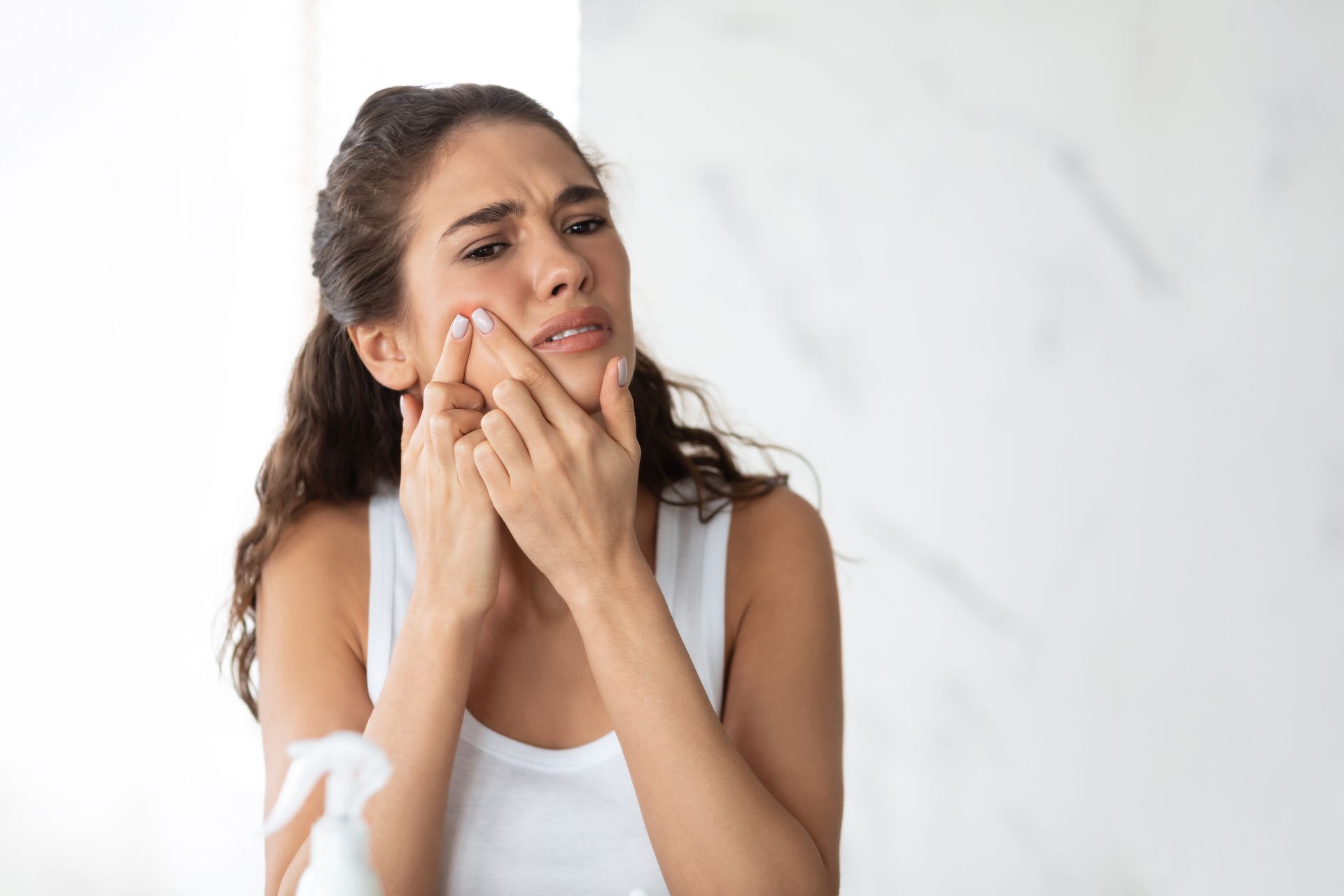 Woman in white tank top looking in mirror, squeezing a pimple on her cheek, concerned expression.