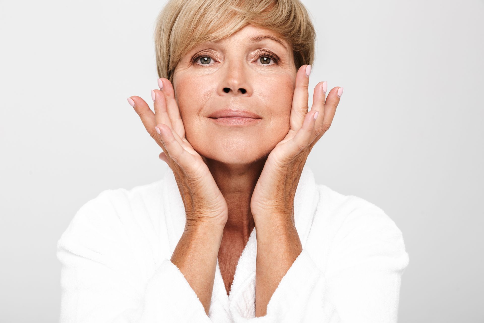 Woman in a white robe, hands on her cheeks, looking upwards, in front of a white background.