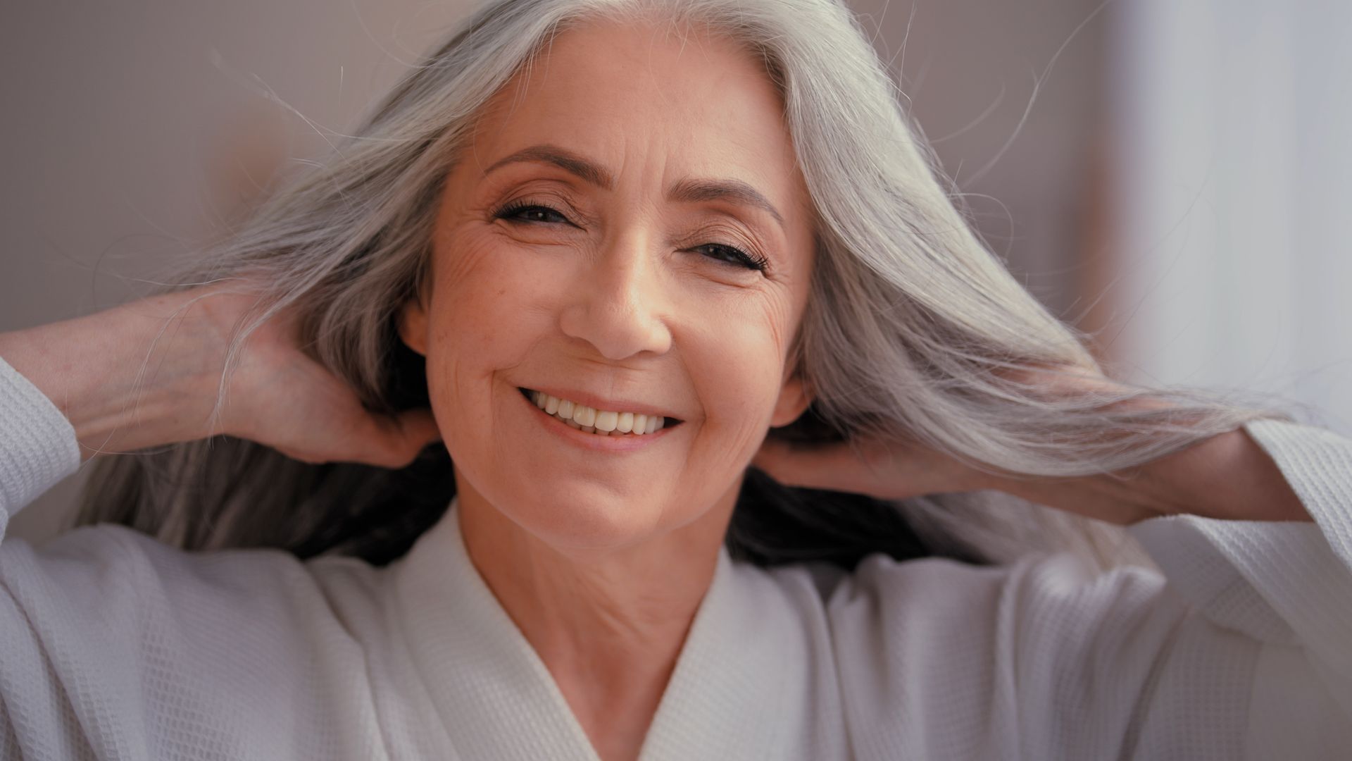 Woman with long silver hair, smiling, holding her hands up, wearing a white robe.