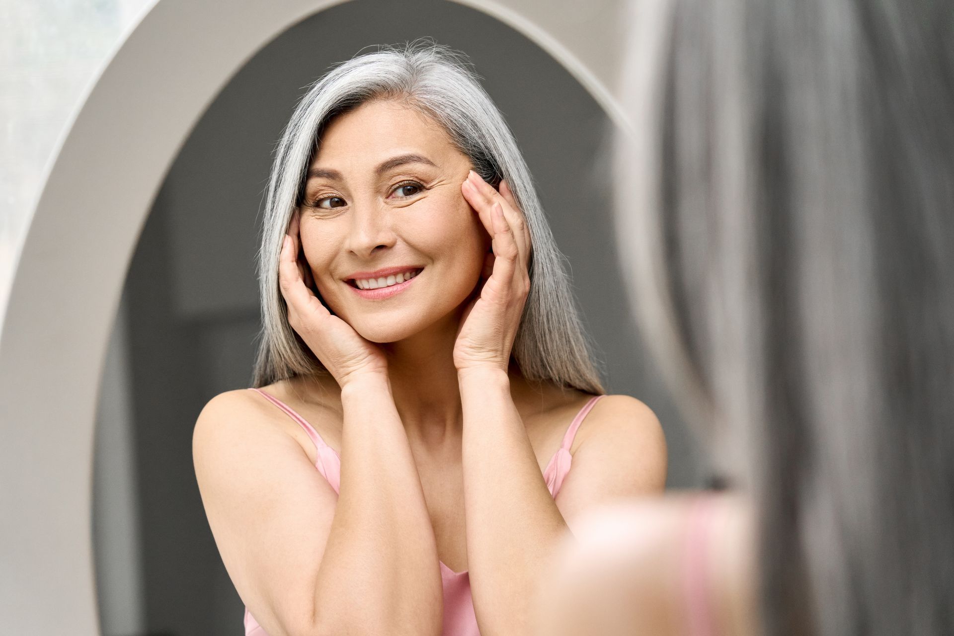 Woman looking in mirror, touching face, smiling. Gray hair, pink top, bathroom setting.