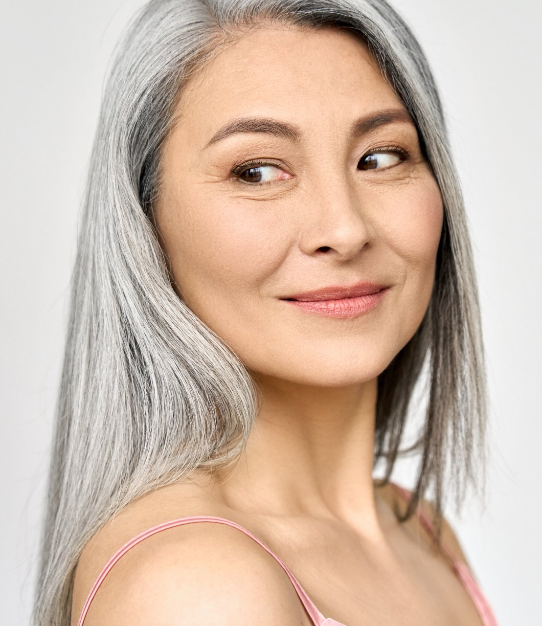Woman with gray hair, looking to the side, smiling slightly, wearing pink top.
