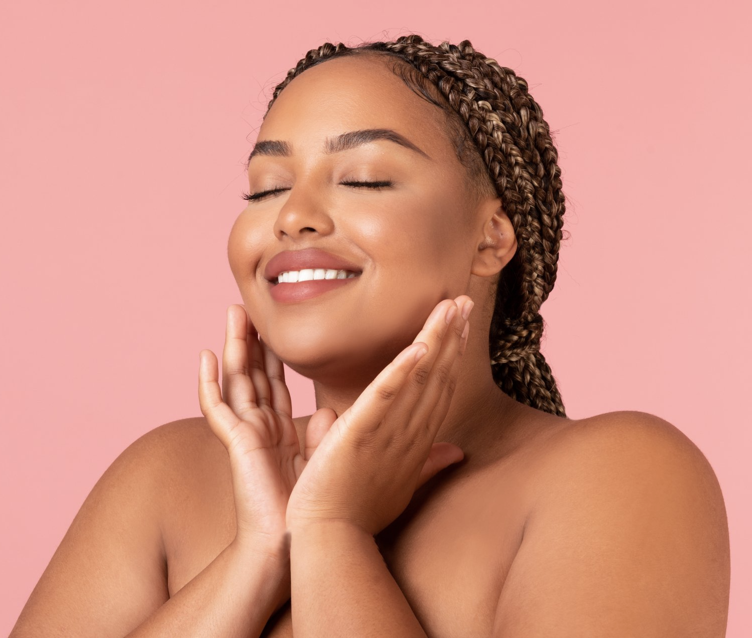 Woman with closed eyes smiling, touching face, against a pink background.