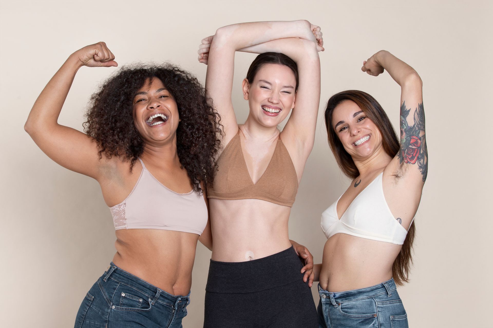 Three women smiling, flexing arms, wearing bras and jeans, tan background.