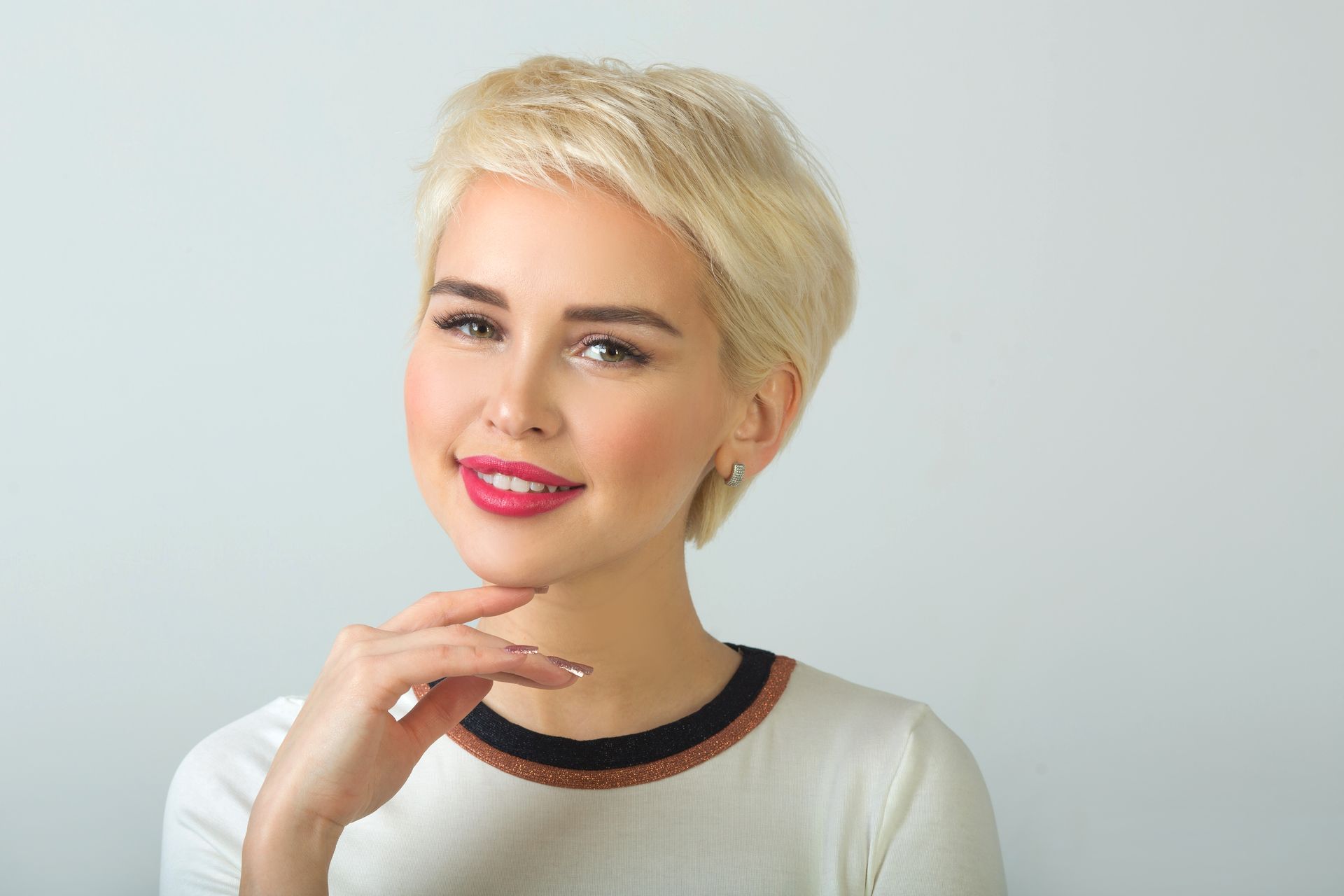 Woman with short blonde hair, smiling, resting chin on hand. She wears a white shirt with a black and brown collar.