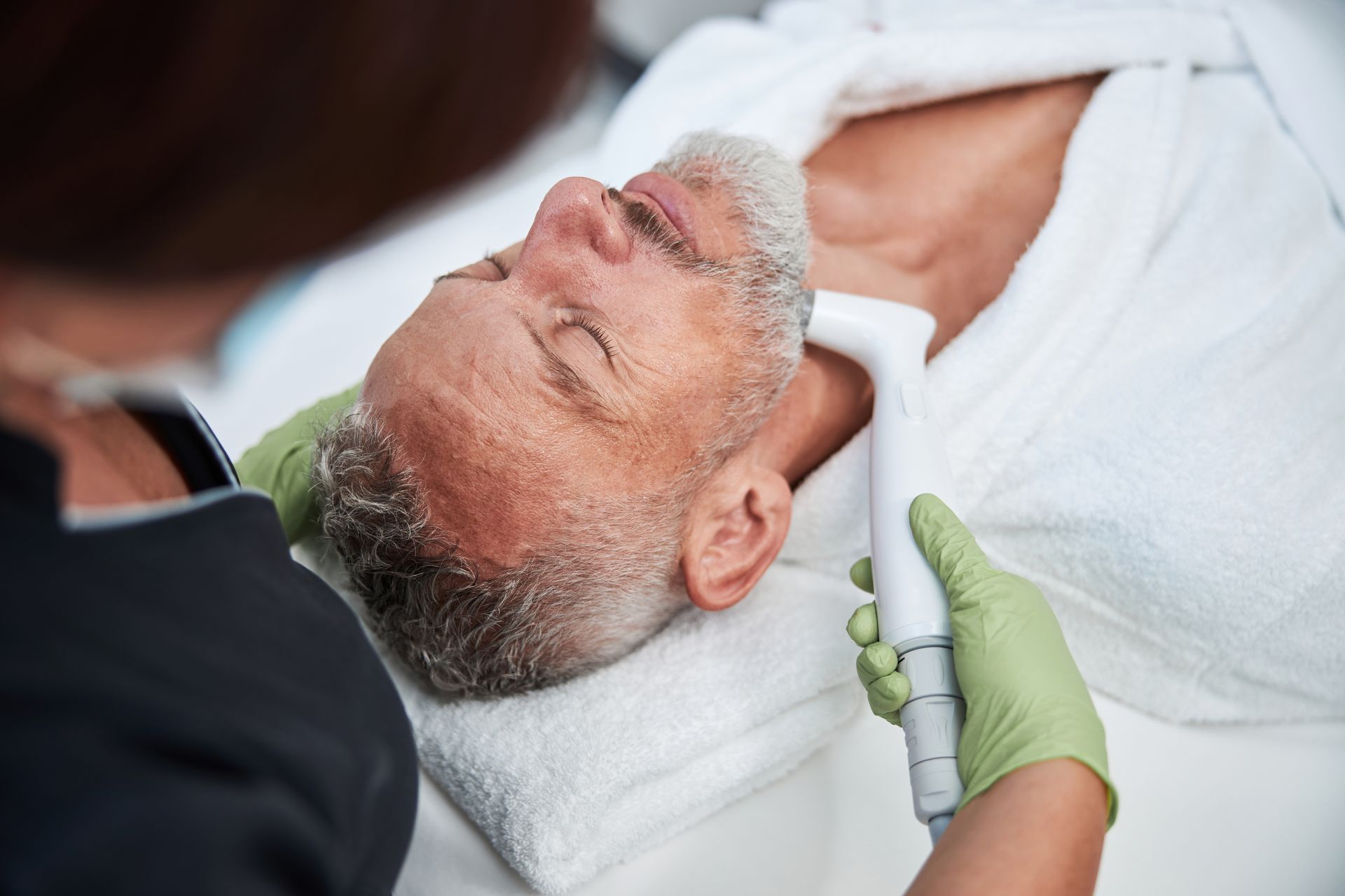 Man receiving neck treatment at a spa. Technician in gloves holds device to his neck. Man reclines on a white towel.