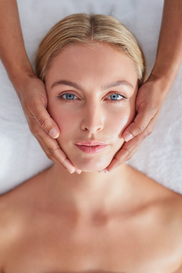 Woman receiving facial massage, face cradled by hands. Blonde hair, blue eyes, on white surface.