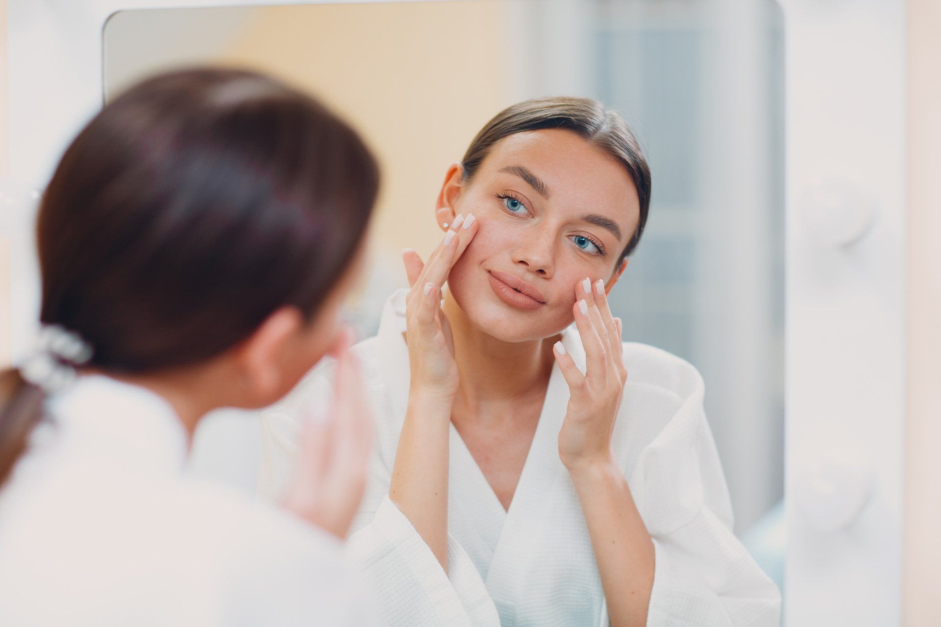 Woman in white robe looking at her face in mirror, applying skincare.