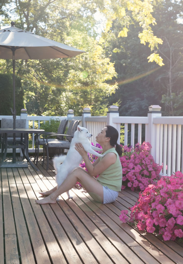 Person holding a white dog on a sunny deck beside pink flowers and a patio table