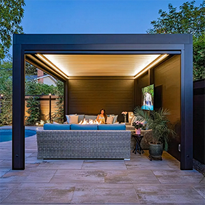 Modern backyard patio cabana with a lit ceiling, sectional sofa, potted plants, and TV at dusk