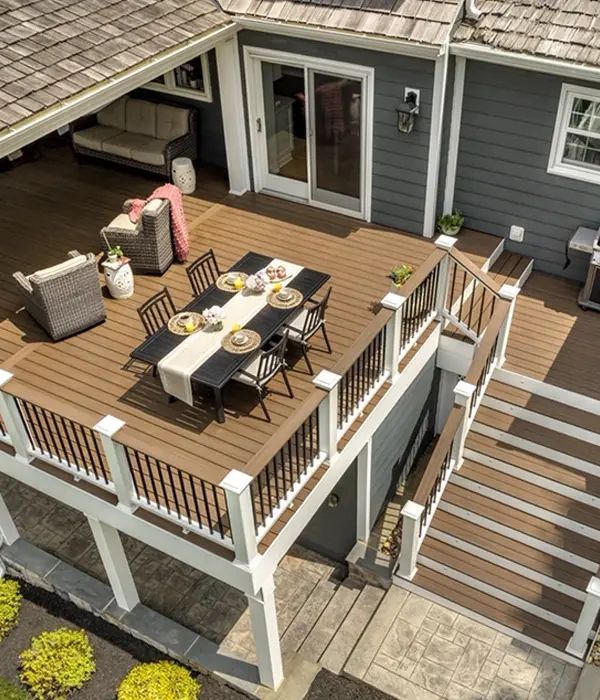 Elevated wooden deck with outdoor dining table, chairs, cushions, and stairs beside a gray house