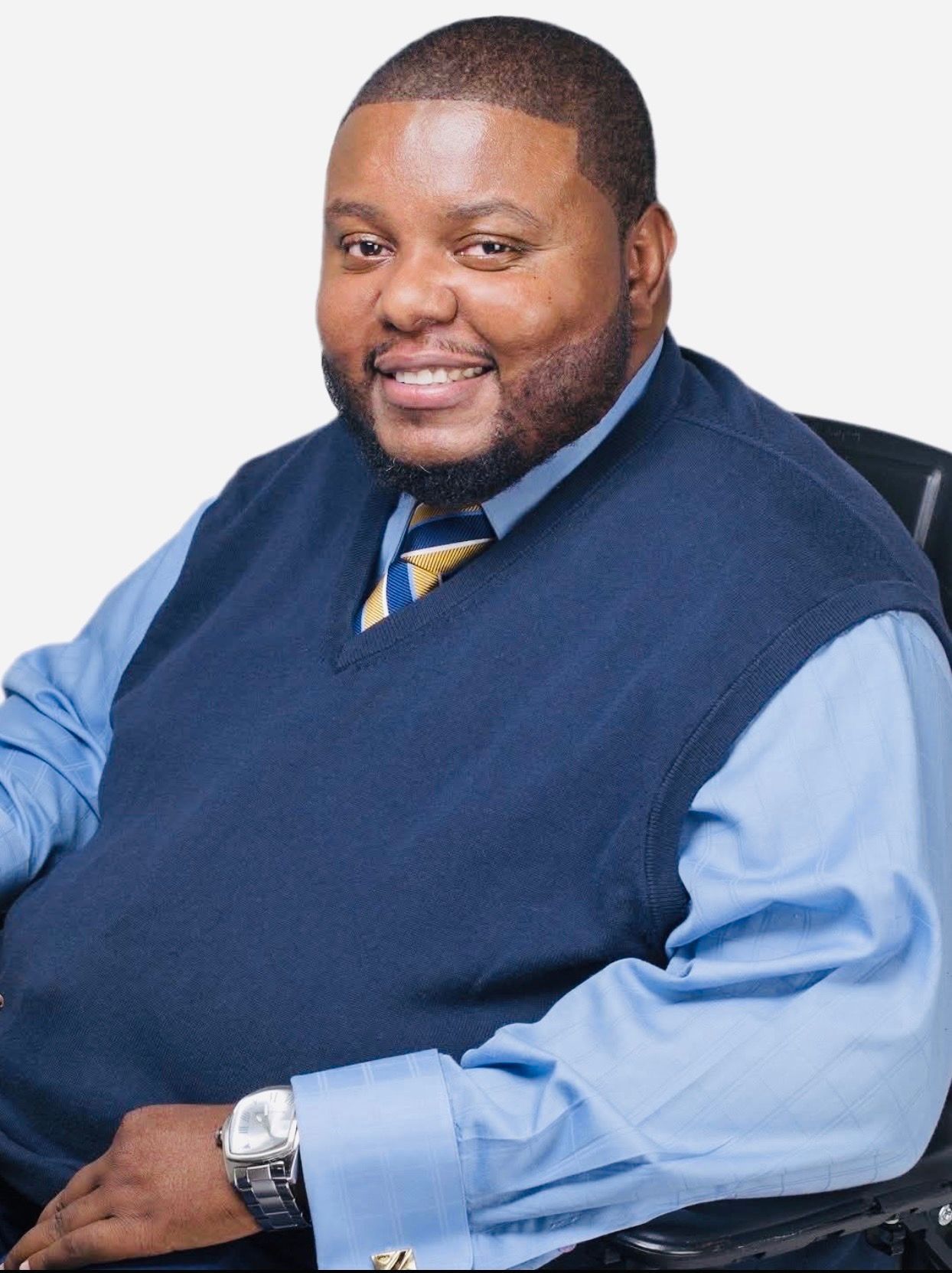 Man in wheelchair smiles near a staircase, wearing a light-colored sweater vest, khakis, and dress shoes.
