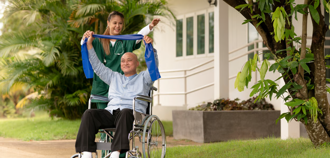 A person in a wheelchair stretches with a blue band, guided by a caregiver. Outdoors.