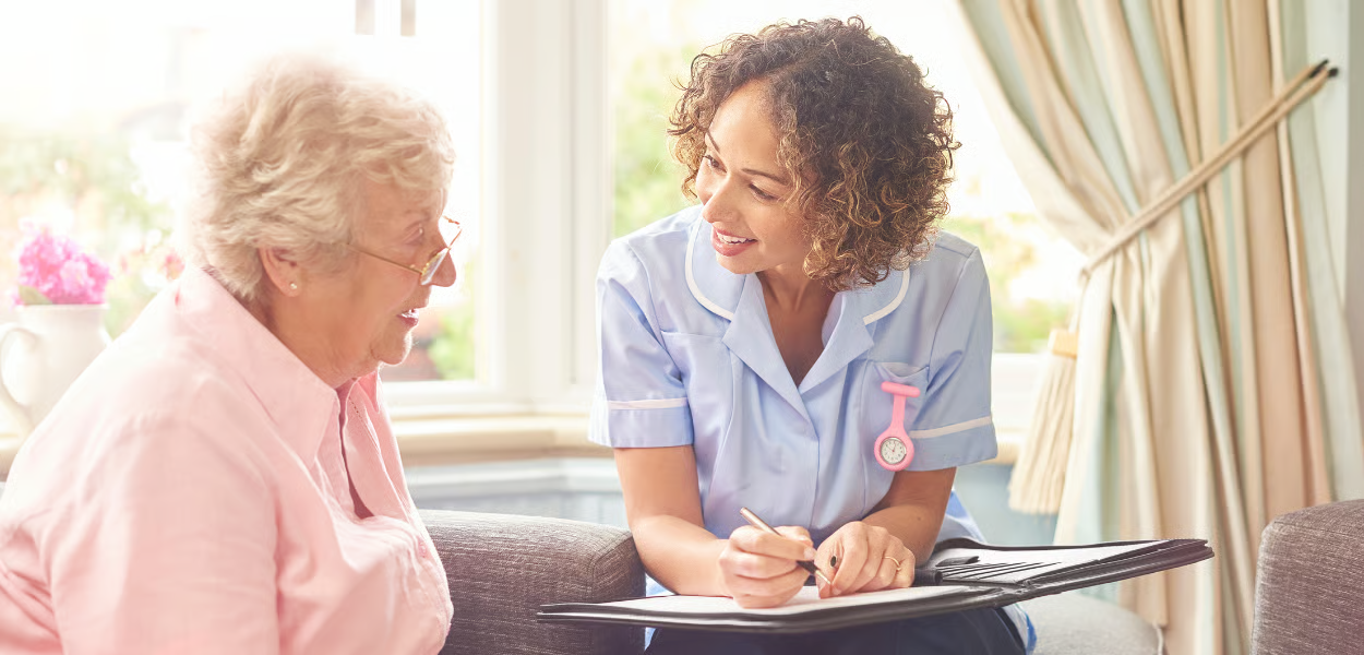 A healthcare worker consults with an elderly person, both seated. They appear to be in a home setting.