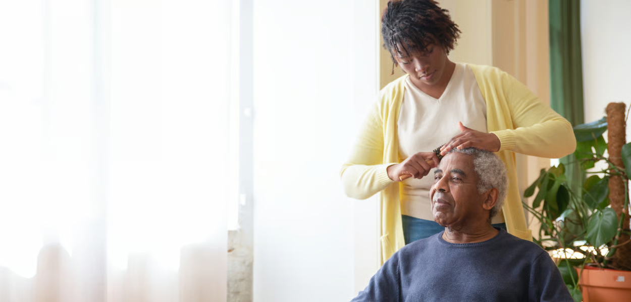 A person combs another person's hair indoors. A plant is in the background.