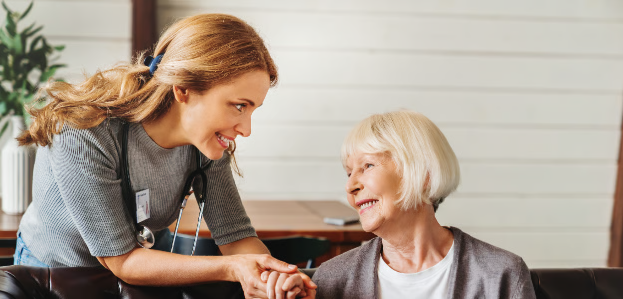 Woman caregiver smiling at elderly woman, holding her hand. Indoor, daytime.