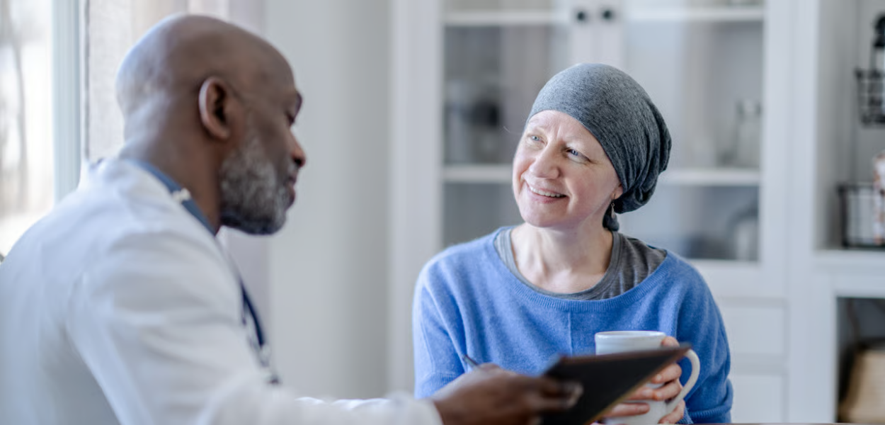 A doctor discusses a tablet with a patient wearing a head covering; the patient holds a mug, and appears concerned.