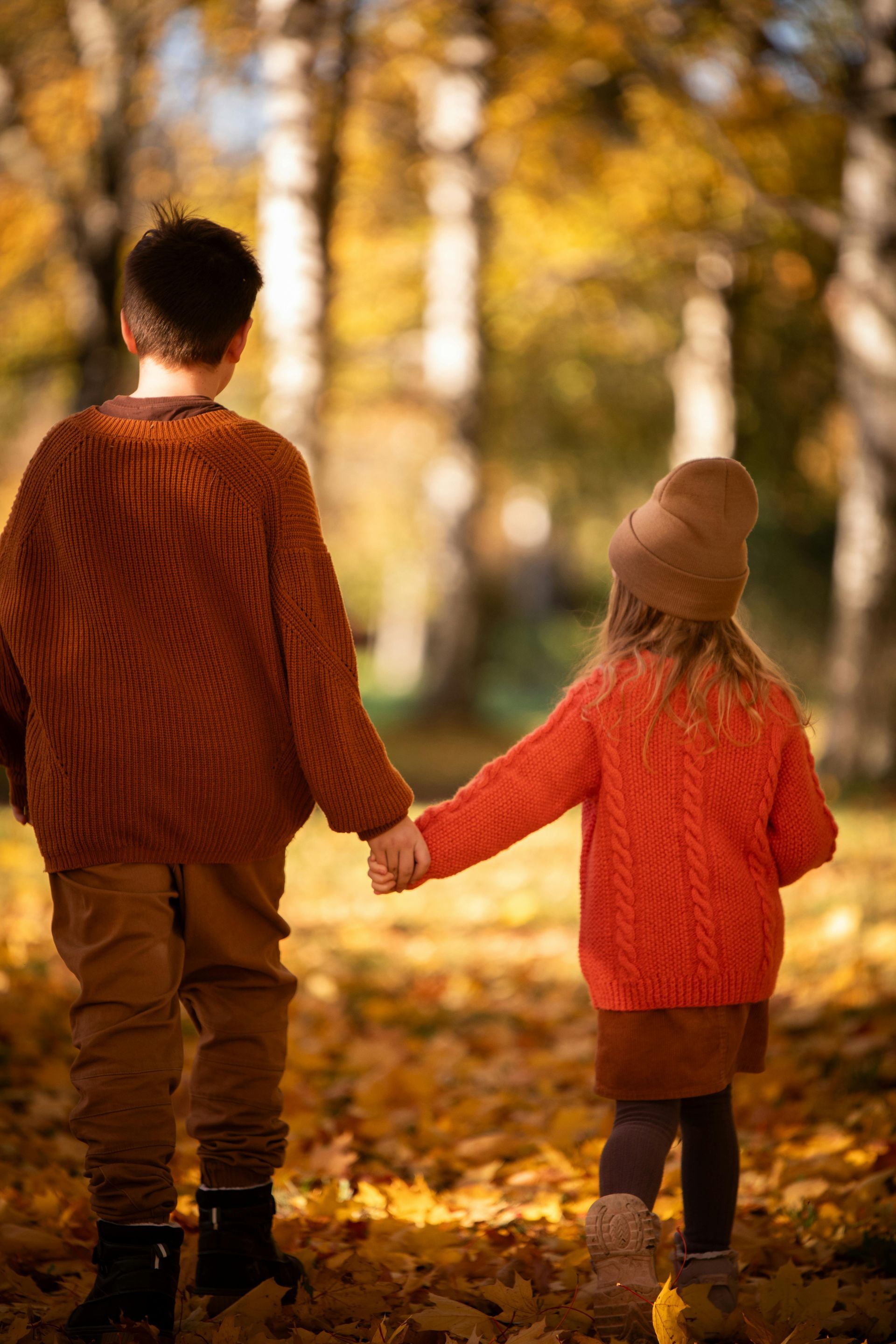 Two children in sweaters hold hands, walking through fall leaves.