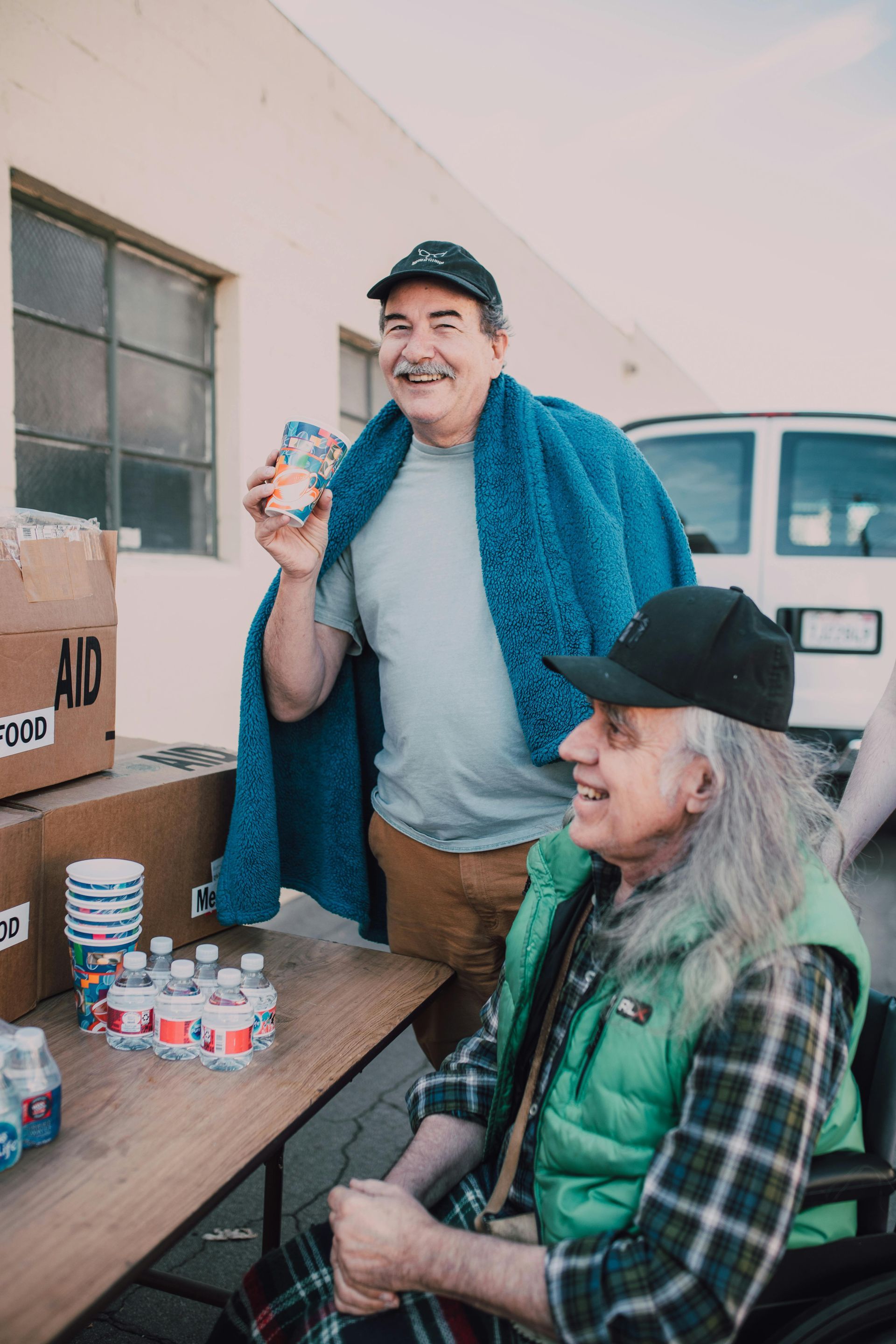 Two men at a food aid station. One holds a drink, the other in a wheelchair, both smiling. Boxes of food and water on table.
