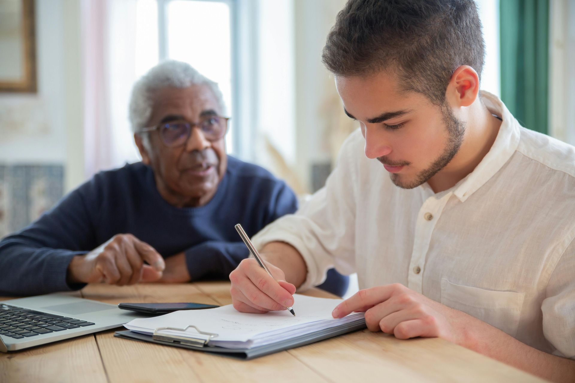 Young person writing on a clipboard, older person watches, sitting at a wooden table.