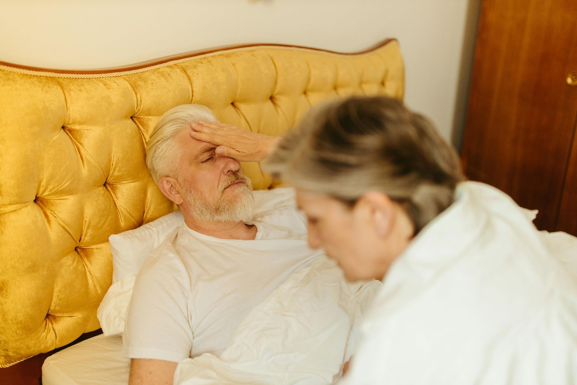 Man in bed, forehead held, appears sick; woman beside him, concerned expression.