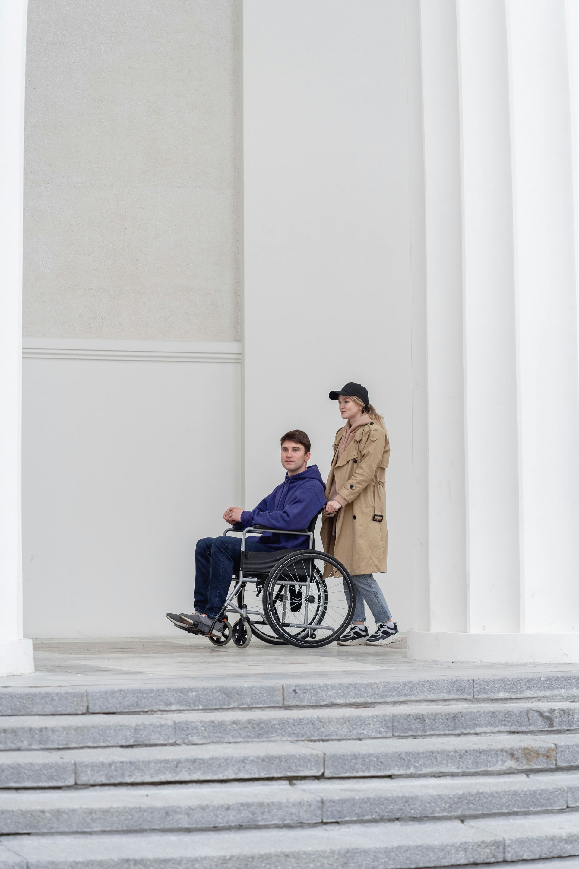 Person in wheelchair being pushed up steps by another person. White columns and building in background.