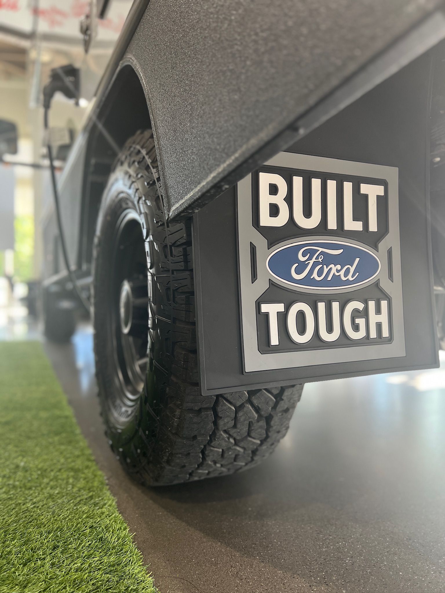 A ford truck with a mudflap that says `` built ford tough '' is parked in a garage.