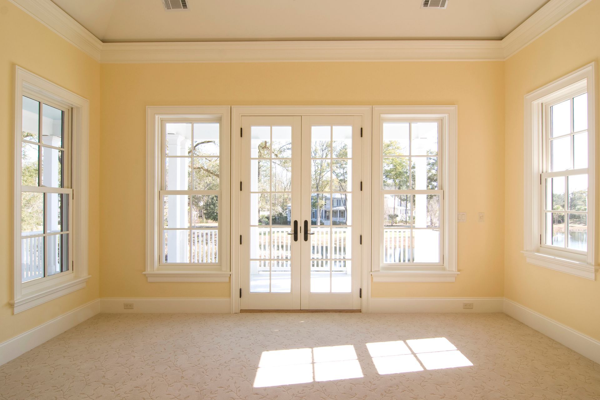 Empty sunlit room with beige carpet, cream walls, and large windows around French doors