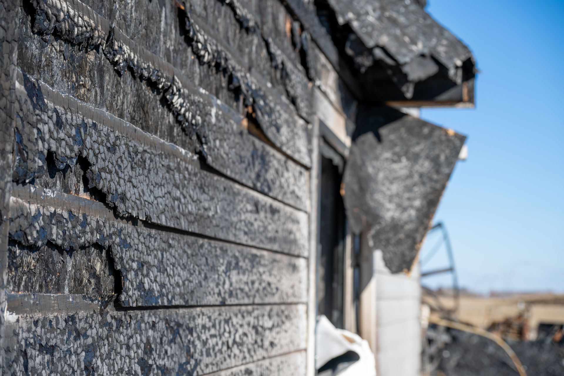 Close-up of charred house siding with heavy fire damage under clear blue sky.