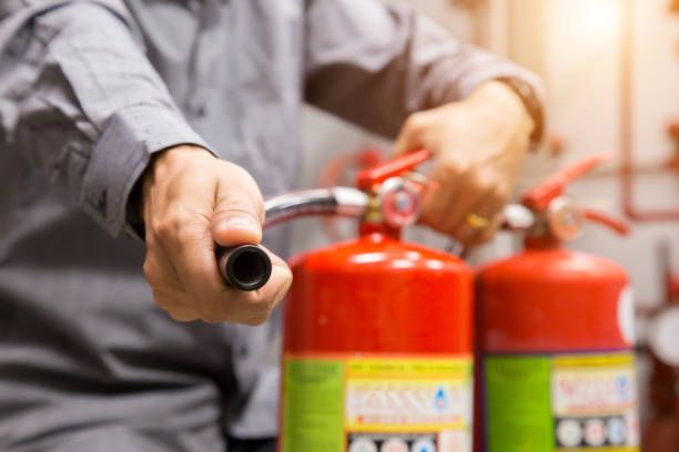Person using fire extinguisher during safety training, holding nozzle on red extinguishers indoors.