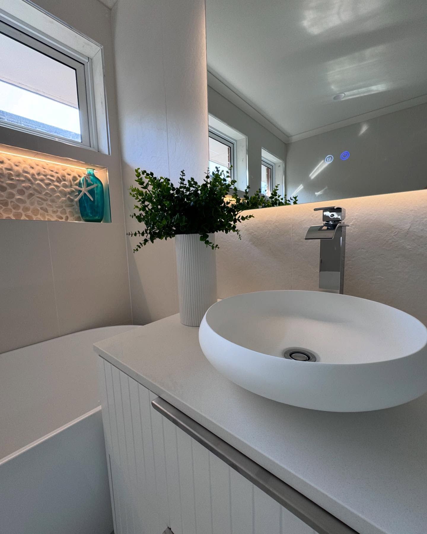 Modern white bathroom vanity with round sink, vase of greenery, and soft natural light