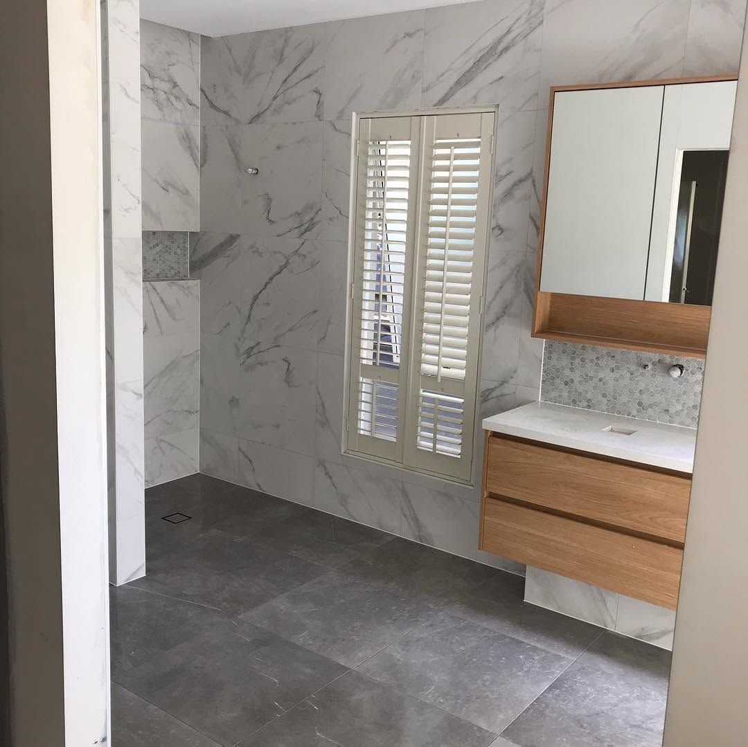Modern bathroom with marble-look walls, gray tile floor, shuttered window, and wood vanity with mirror.