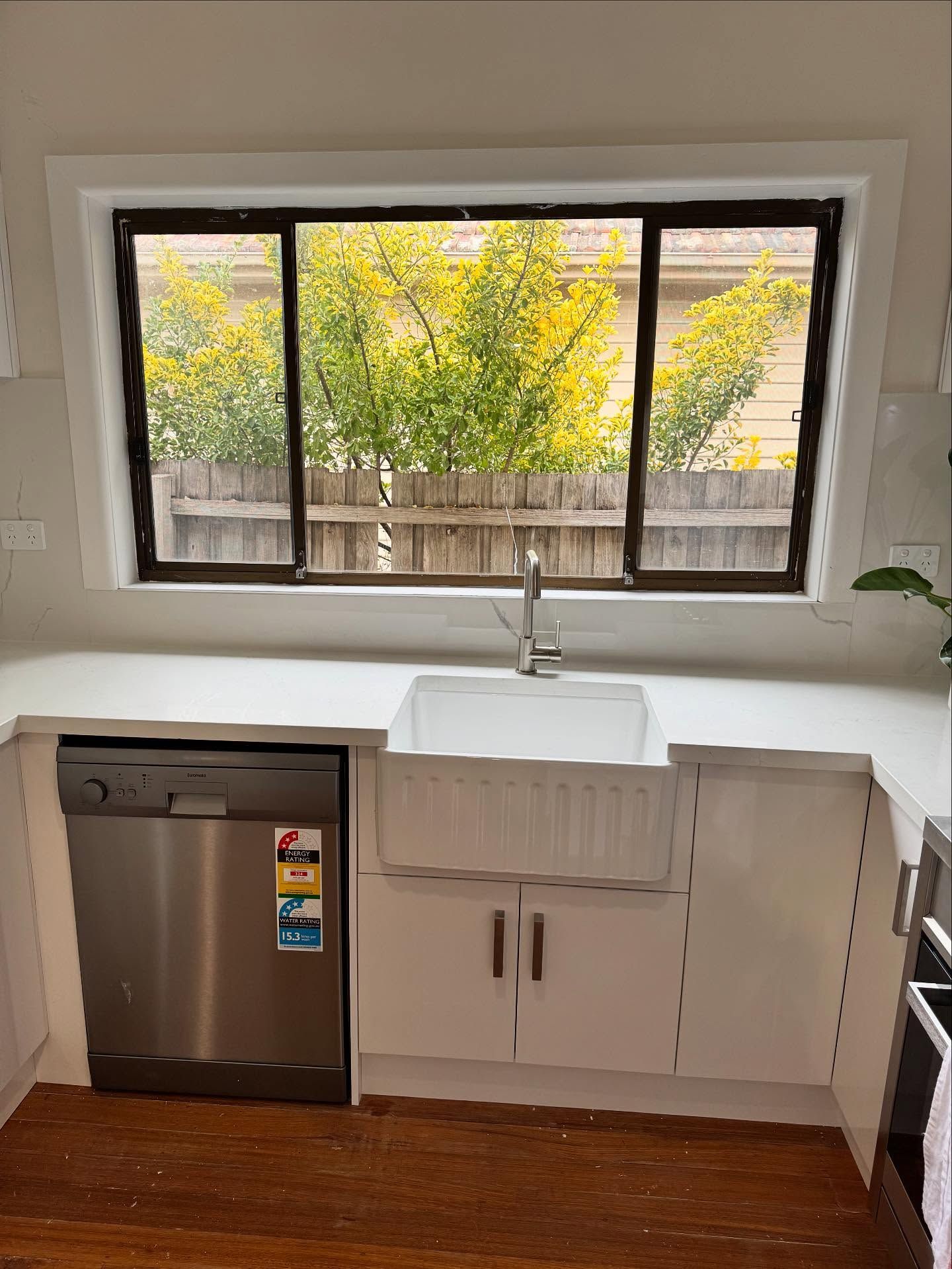 Compact kitchen sink under a window with stainless dishwasher and white cabinets, overlooking yellow trees.